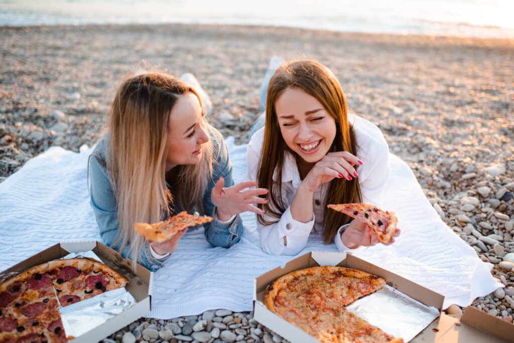 Two young beautiful girlfriends having fun with pizza at beach over sea shore outdoors in sun light. Summer vacation season. Friendship. Happiness. Sisters spend time together at coast line.